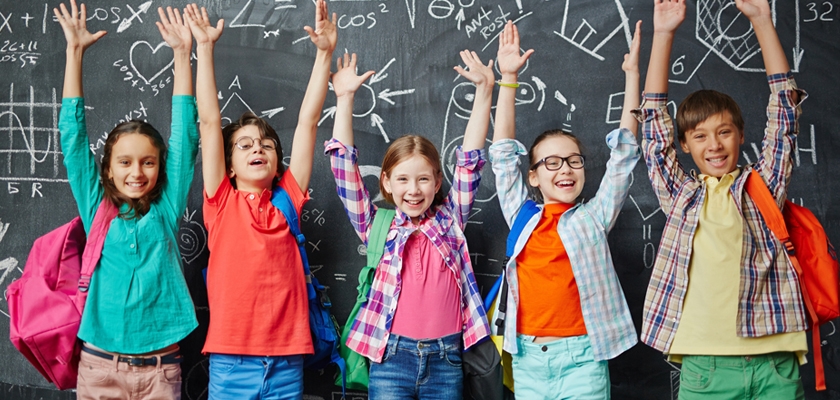 Five cheerful school children raising their hands in excitement, in front of a chalkboard.