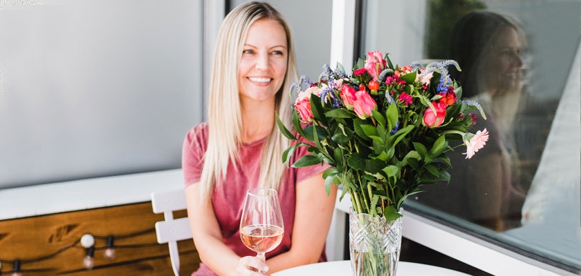 A woman enjoying a glass of rosé wine, sitting by a vase of vibrant flowers.