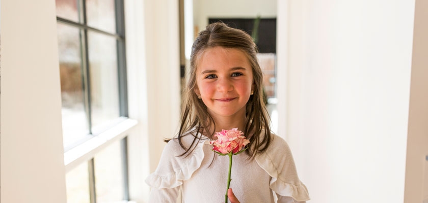 A smiling girl holding a pink flower, radiating joy in a bright hallway.