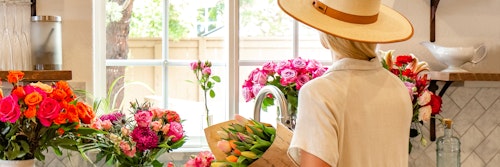 Woman in a beige hat arranging colorful bouquets of roses and tulips in a bright, sunny kitchen interior with open windows and stylish decor.