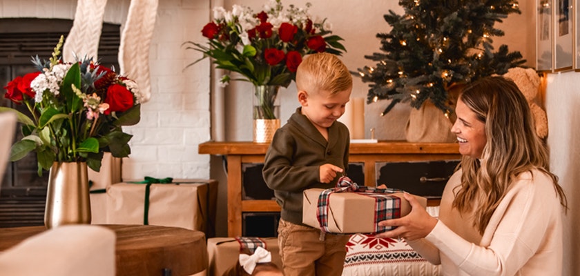 A joyful child presenting a beautifully wrapped gift to a smiling woman beside festive decorations.