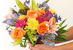 A person holding a vibrant bouquet of flowers including orange roses, pink lilies, yellow blooms, and various purple flowers against a white background.