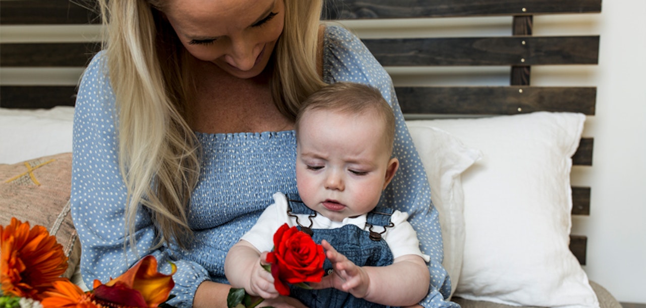 Woman in a blue top smiling at a baby holding a red rose, with orange flowers nearby, portraying a tender moment between mother and child at home.