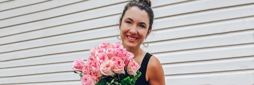 Smiling woman with a bun holding a large bouquet of pink roses against a white horizontal siding background, exuding a joyful and warm ambiance.