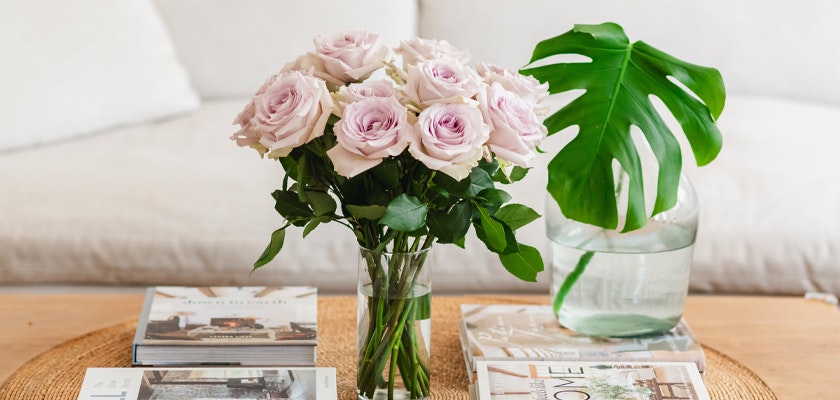 Bouquet of pale pink roses in a glass vase on a coffee table with magazines and a monstera leaf in a vase in a cozy living room setting.