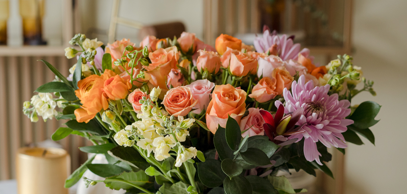 Bright floral arrangement featuring peach and pink roses with lush greenery and daisies.