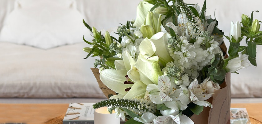 Elegant bouquet of white lilies and assorted greenery wrapped in brown paper on a wooden table, with a cozy bedroom setting featuring pillows in the background.