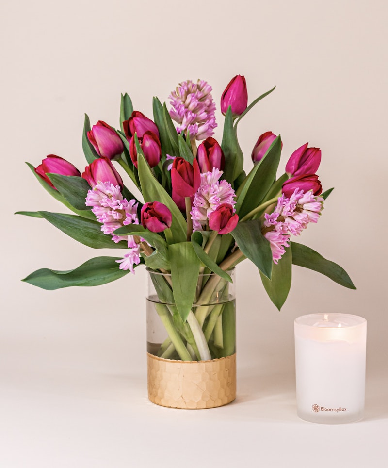 A vibrant bouquet of pink tulips and purple hyacinth flowers in a clear glass vase with a gold accent, accompanied by a lit white candle on a neutral background.