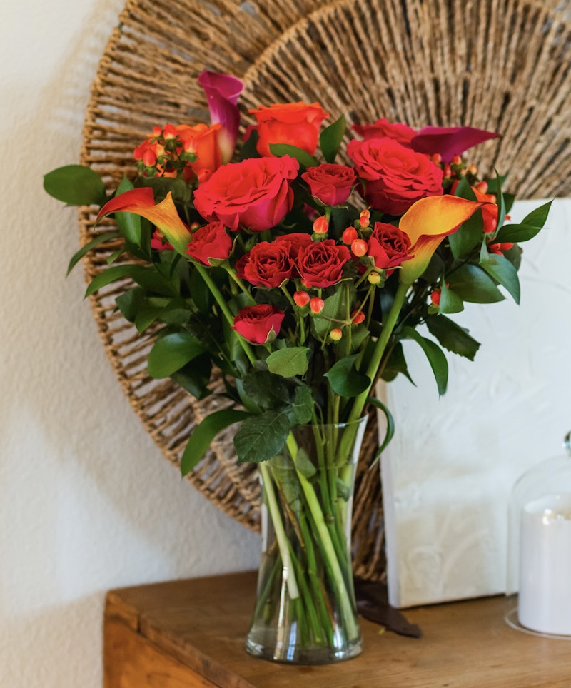 Vibrant bouquet of red roses and orange lilies with green foliage in a clear vase against a rustic wicker background on a wooden table, adding a warm touch to home decor.