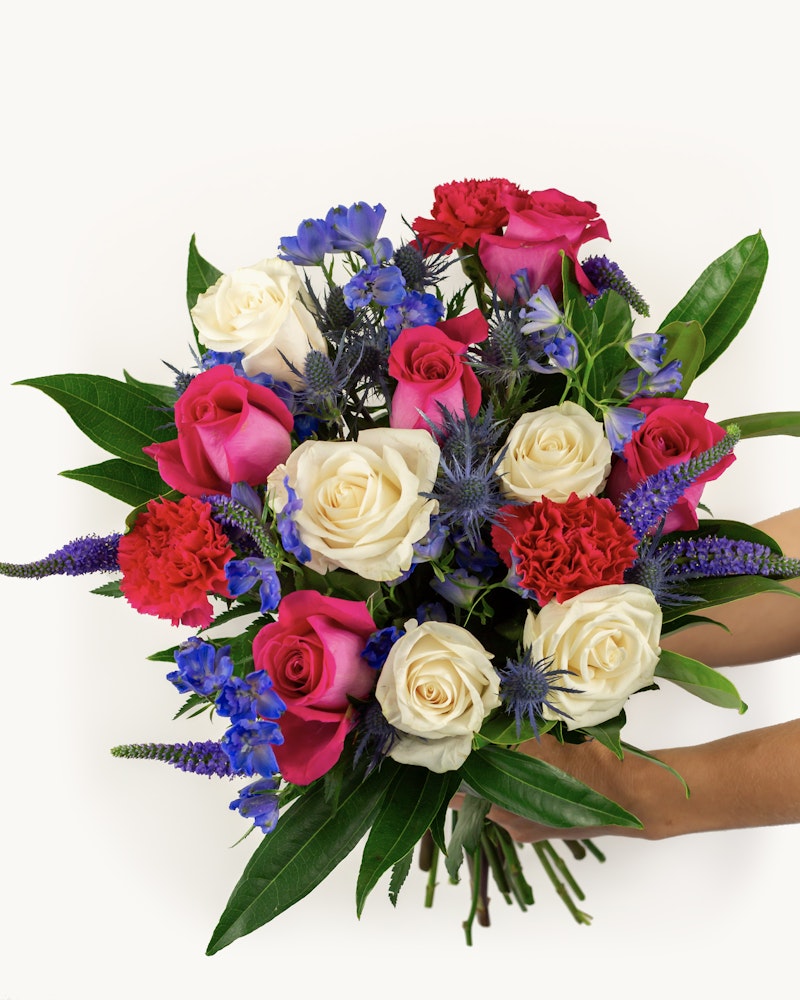 A vibrant bouquet with a mix of pink and white roses, blue flowers, and lush greenery held against a white background, showcasing a range of colors and textures.