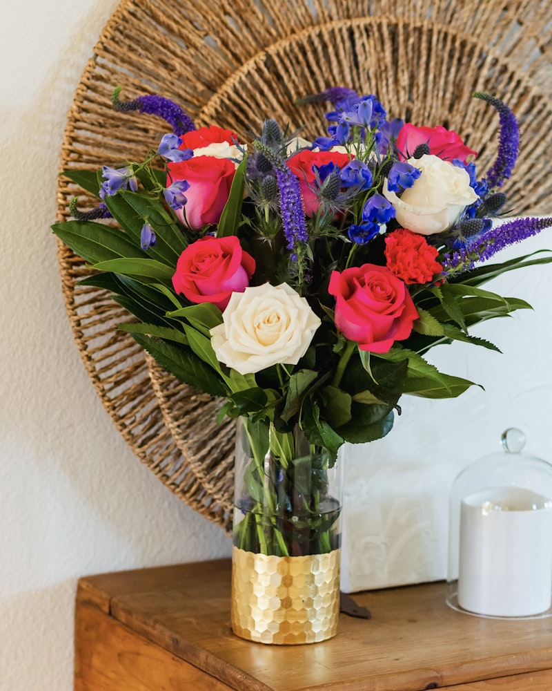 Beautiful bouquet of colorful flowers with red, pink, and white roses, blue accents, and green foliage in a gold-detailed vase on a wooden table beside a white candle.