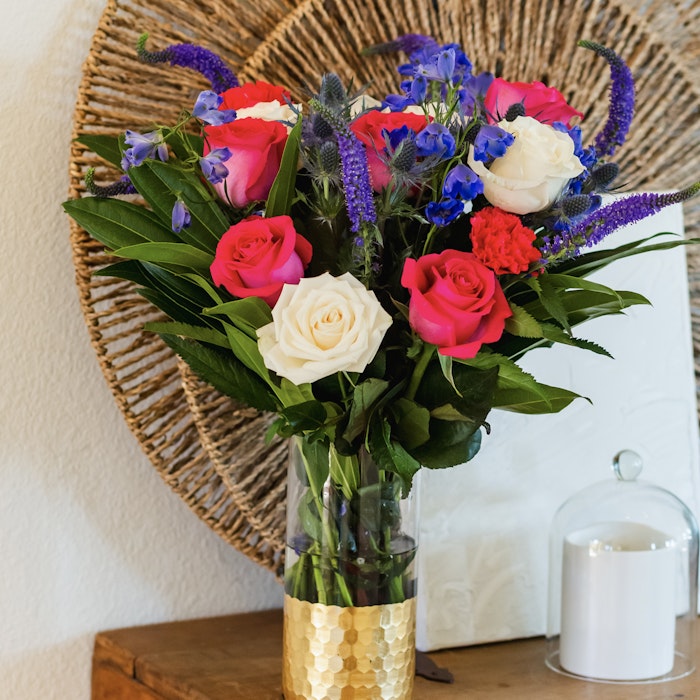 Beautiful bouquet of colorful flowers with red, pink, and white roses, blue accents, and green foliage in a gold-detailed vase on a wooden table beside a white candle.