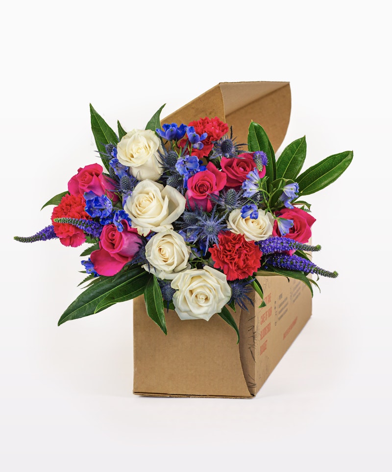 Vibrant bouquet of flowers with white roses, red carnations, and blue accents presented in a brown cardboard box on a white background, perfect for gifting.