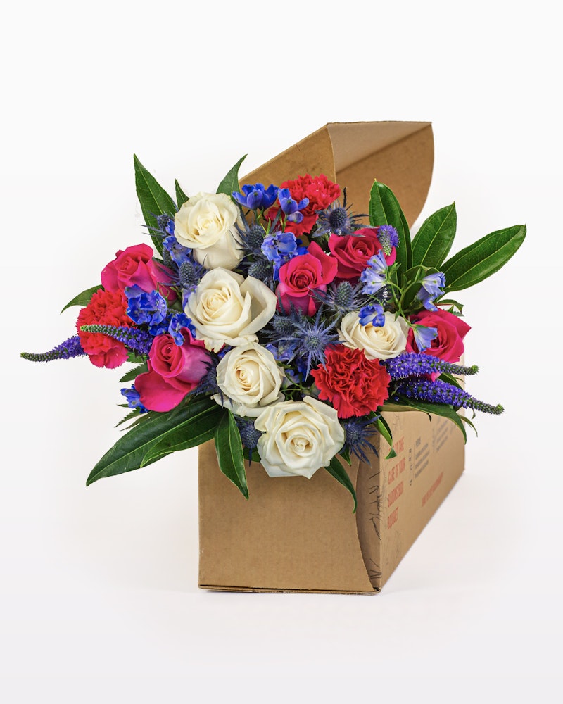 Vibrant bouquet of flowers with white roses, red carnations, and blue accents presented in a brown cardboard box on a white background, perfect for gifting.