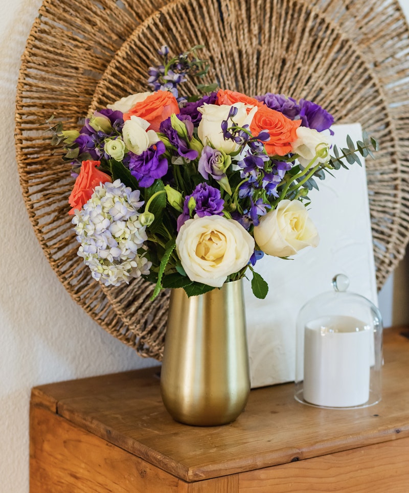 Vibrant bouquet of flowers with white roses, purple accents, and orange blossoms in a golden vase, set against a wicker background on a wooden surface.