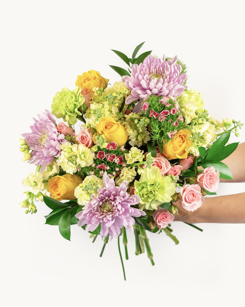 A vibrant bouquet of fresh flowers including yellow roses, pink chrysanthemums, and green accents cradled in a person's hands against a white background.