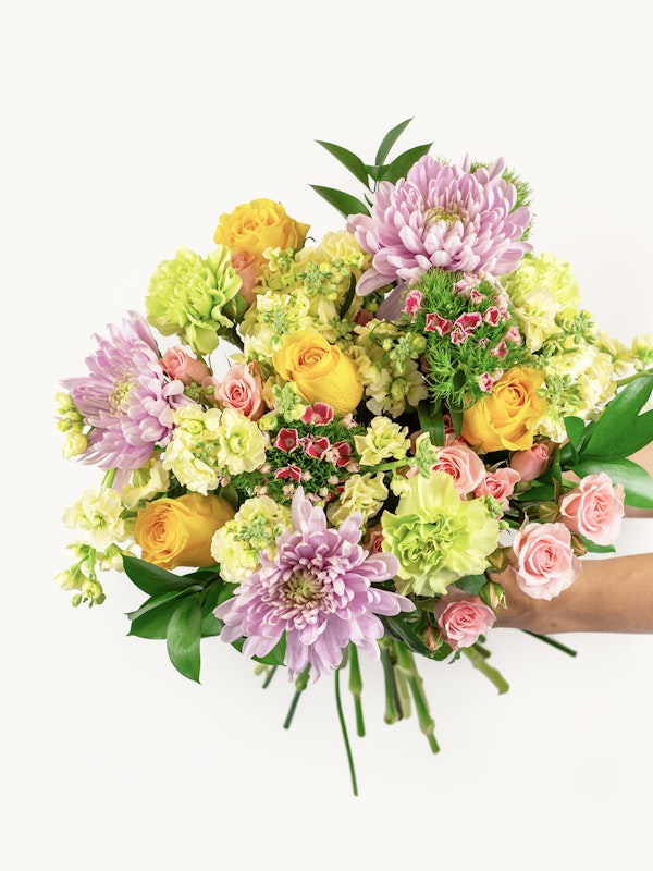 A vibrant bouquet of fresh flowers including yellow roses, pink chrysanthemums, and green accents cradled in a person's hands against a white background.