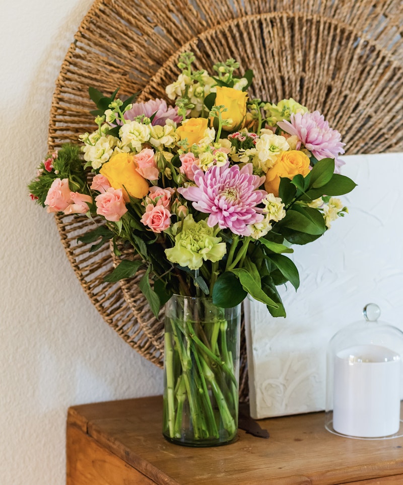 Vibrant bouquet of yellow roses, pink flowers, and purple blooms in a clear glass vase on a wooden table, with a wicker wall decoration and white candle in the background.