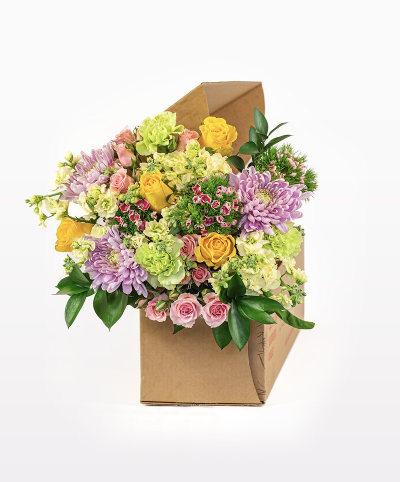 Bright and vibrant bouquet of mixed flowers including yellow roses, pink roses, purple chrysanthemums, and greenery in a cardboard gift box on a white background.