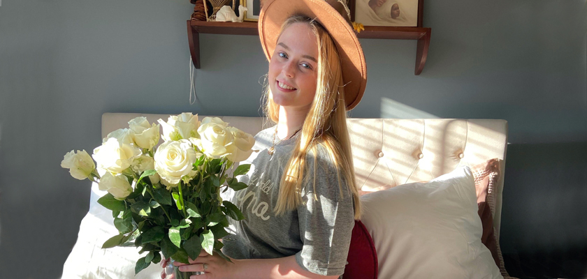 Young woman sitting on a bed, smiling while holding a bouquet of white roses, wearing a stylish hat.