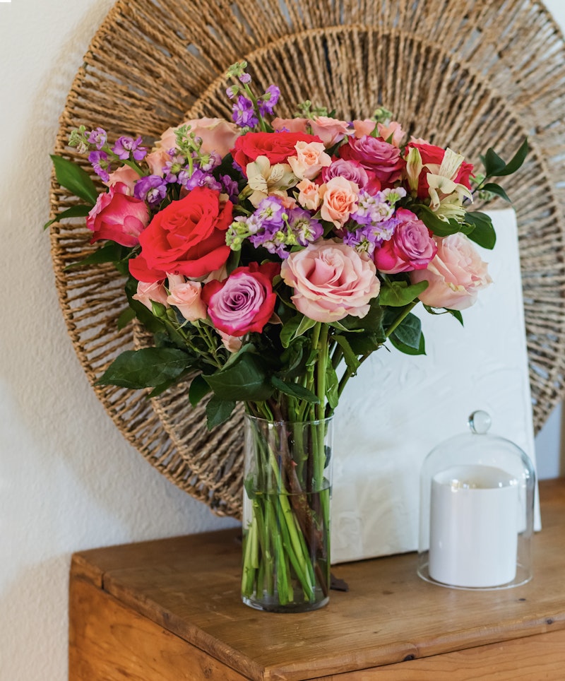 Vibrant bouquet of pink and red roses with purple flowers in a clear vase on a wooden table against a woven circular backdrop, with a white candle lantern alongside.