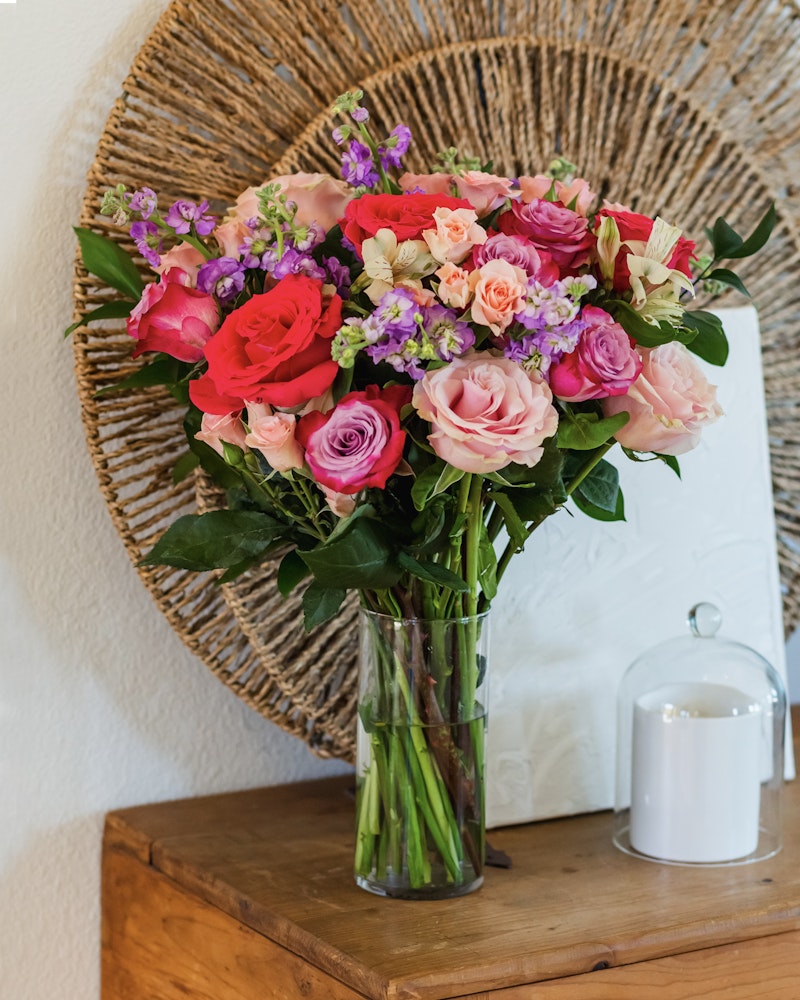 Vibrant bouquet of pink and red roses with purple flowers in a clear vase on a wooden table against a woven circular backdrop, with a white candle lantern alongside.
