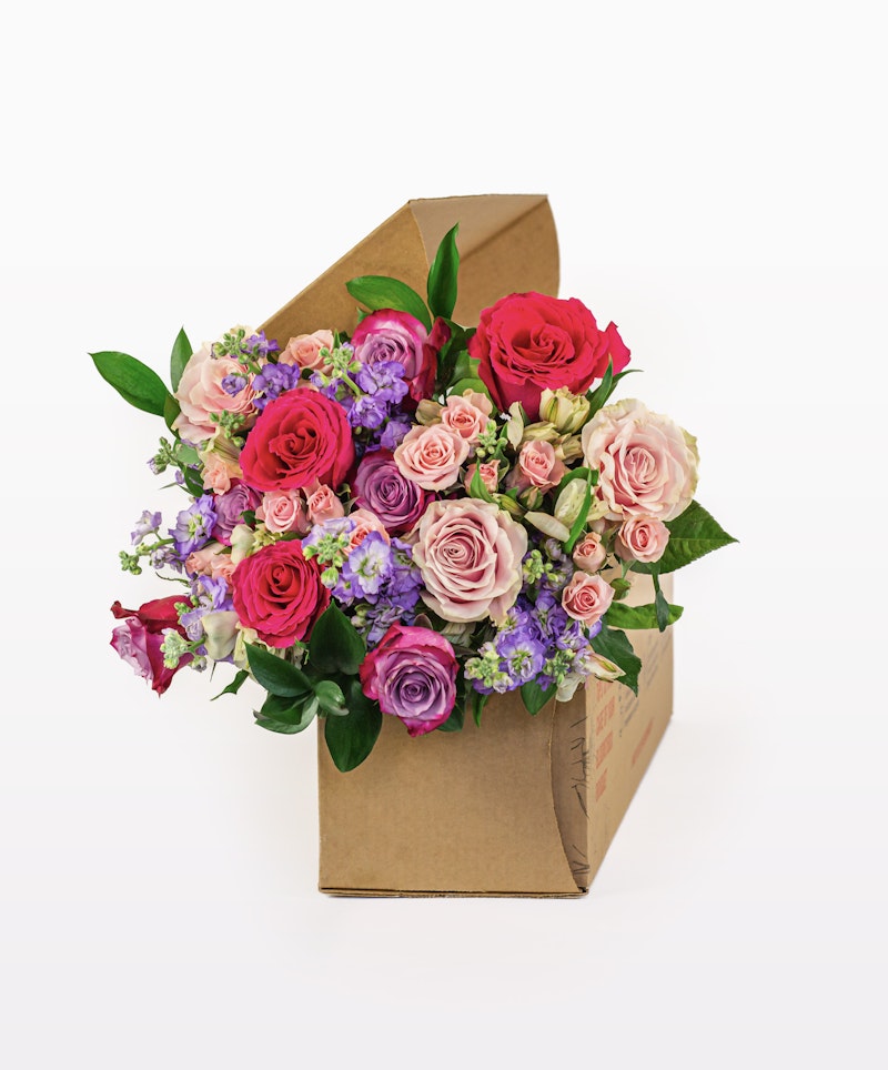 An arrangement of vibrant red, pink, and purple flowers with green leaves, presented in a stylish brown cardboard gift box against a white background.