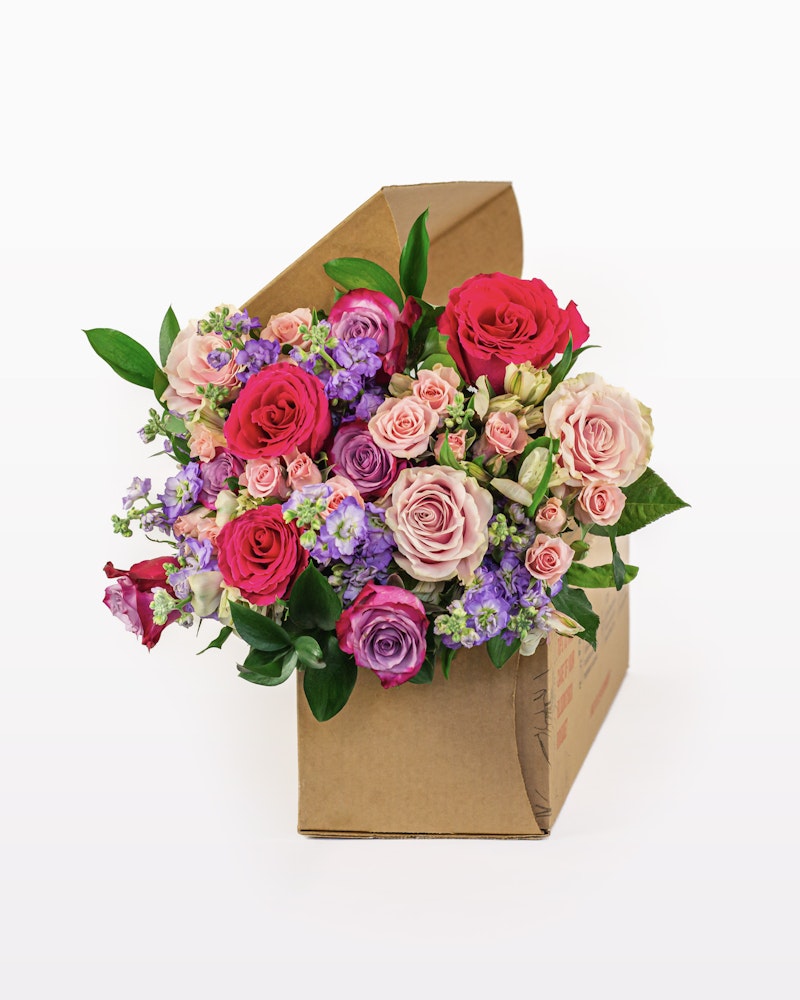 An arrangement of vibrant red, pink, and purple flowers with green leaves, presented in a stylish brown cardboard gift box against a white background.