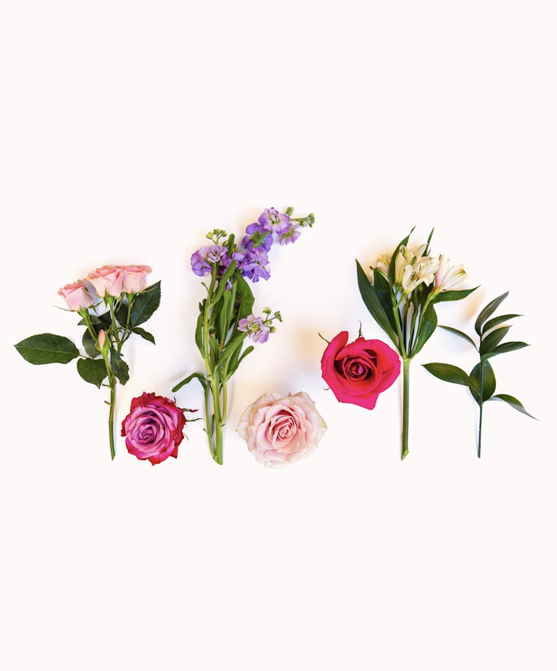 Assorted fresh flowers including pink and red roses, purple blooms, and white lilies arranged in a neat row on a clean, white backdrop.