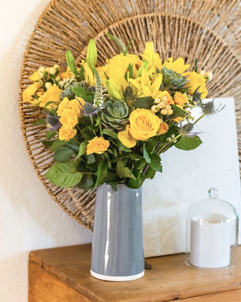 Bright yellow and orange flowers arranged in a modern gray vase on a wooden table with decorative items and a woven wall art piece in the background.