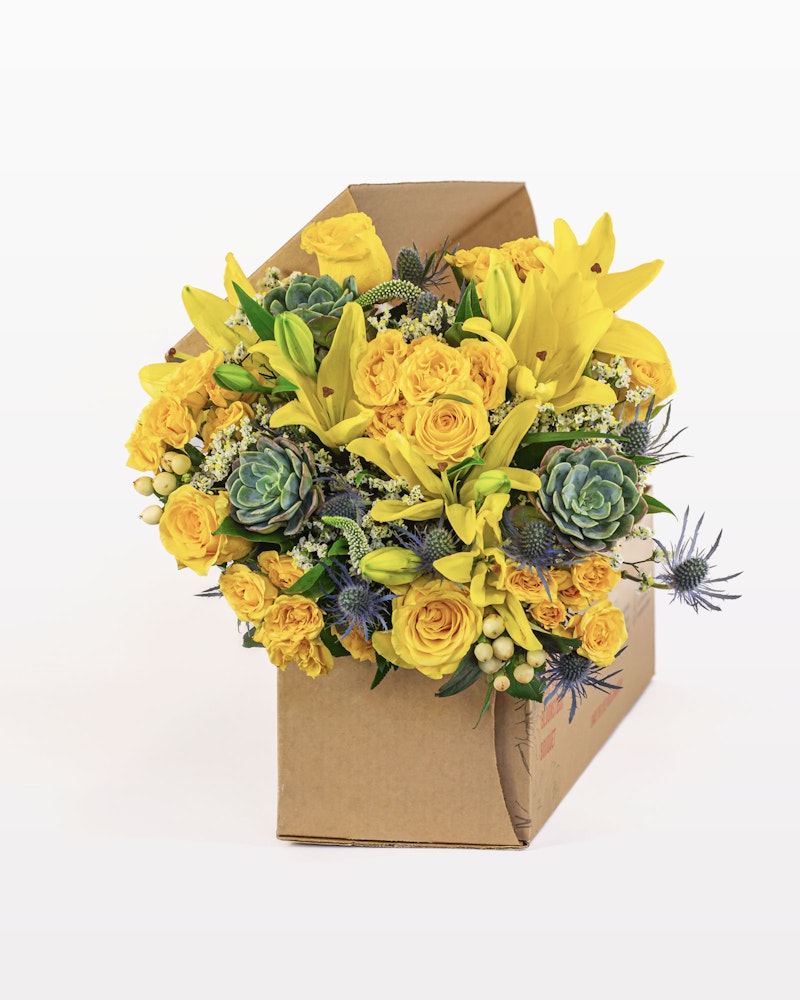 Vibrant bouquet of yellow roses, lilies, and green succulents with hints of blue flowers, elegantly arranged in a brown cardboard box against a white background.