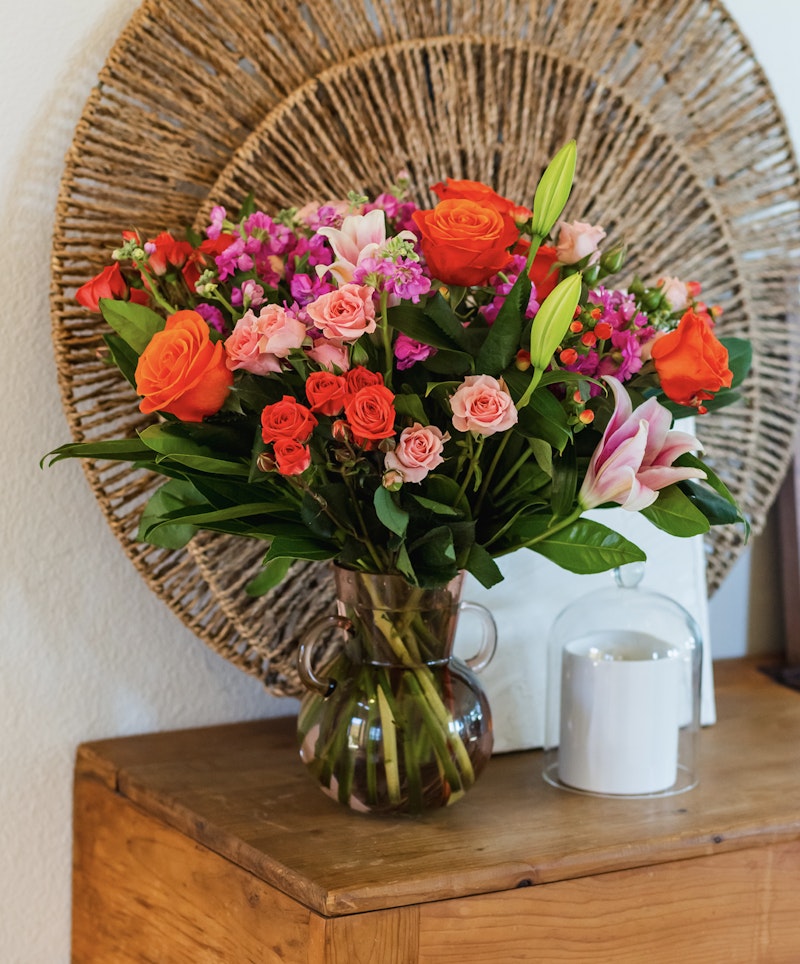 Vibrant bouquet of orange roses, pink blooms, and lilies in a glass vase on a wooden table, with a decorative wicker plate hanging in the background.