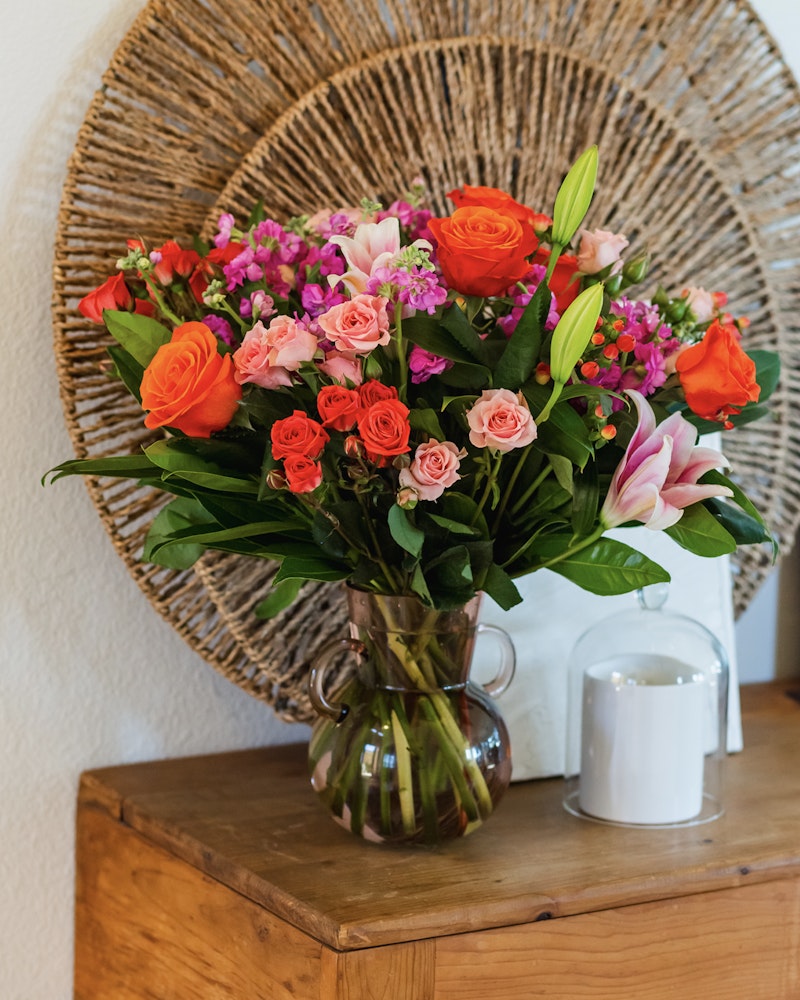 Vibrant bouquet of orange roses, pink blooms, and lilies in a glass vase on a wooden table, with a decorative wicker plate hanging in the background.