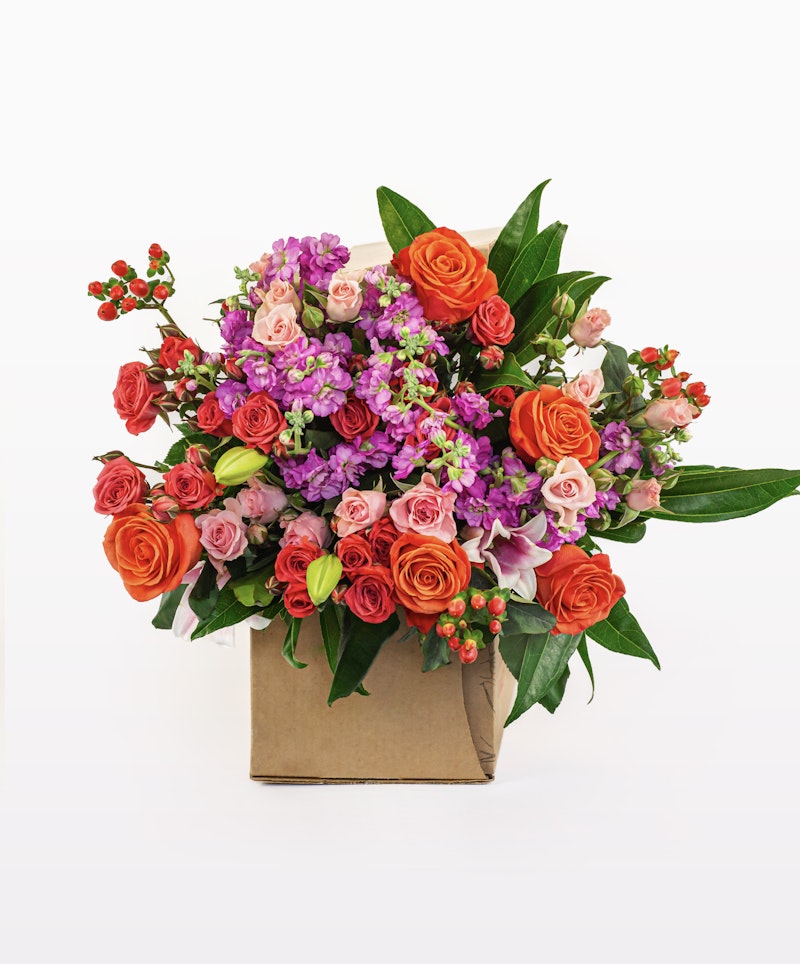 Vibrant bouquet of fresh flowers including orange roses, pink roses, purple accents, and green leaves packaged in a stylish brown paper bag on a white background.