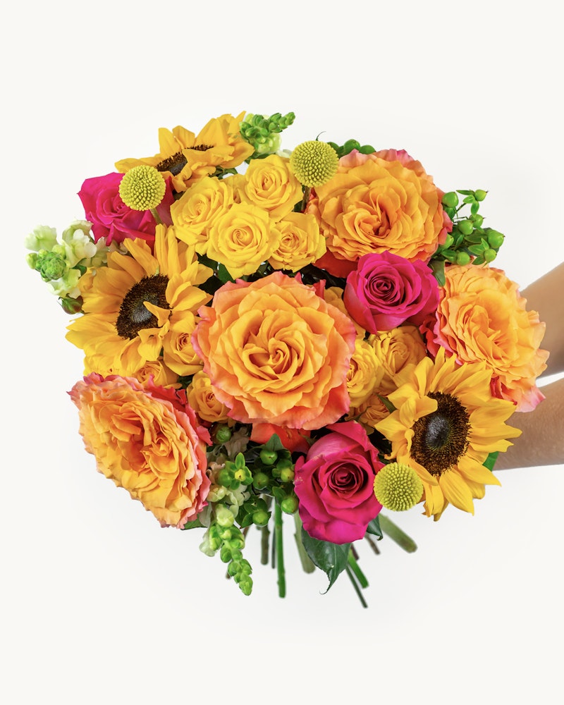 Vibrant bouquet of flowers featuring orange roses, yellow sunflowers, pink blooms, and green accents held in hands against a white background.