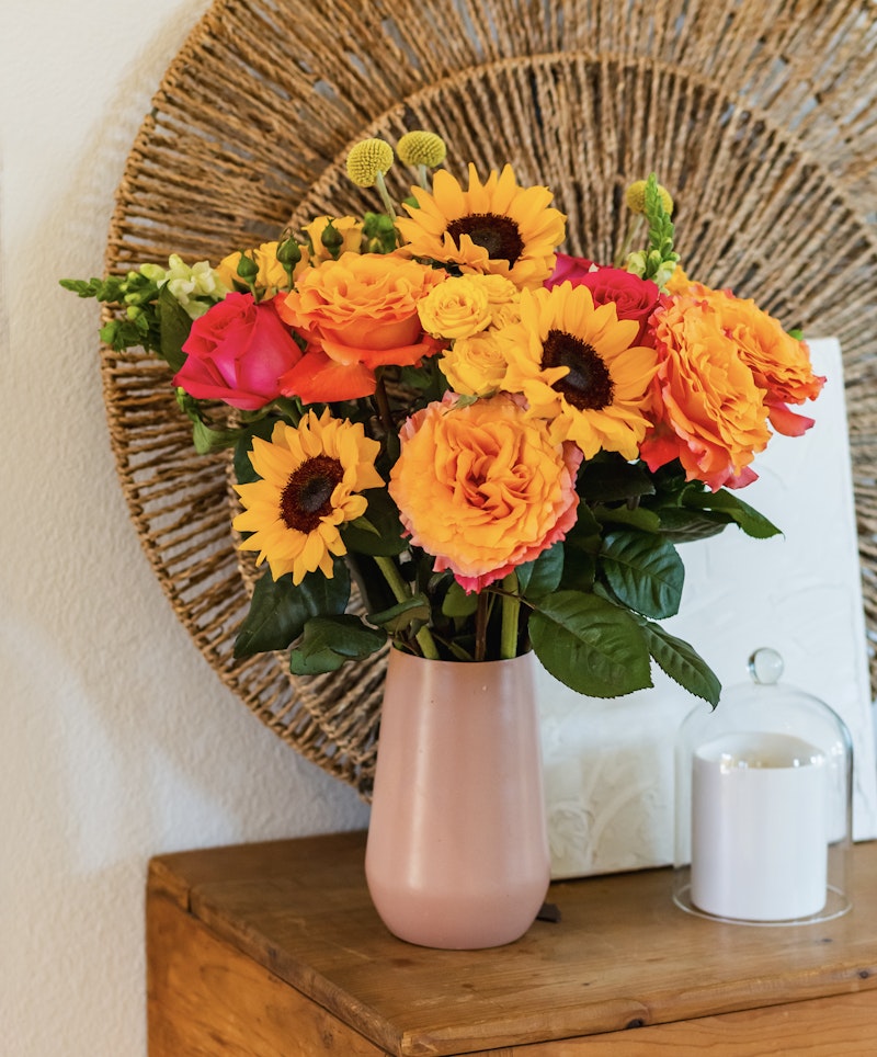 Vibrant bouquet of orange roses and sunflowers in a pink vase on a wooden table, with a round woven wall decoration and a white candle in the background.