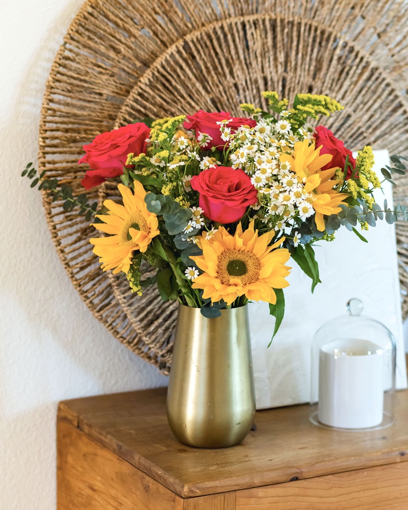 Vibrant floral arrangement featuring red roses and yellow sunflowers in a gold vase on a wooden table against a straw wall decor backdrop, with a white candle lantern nearby.