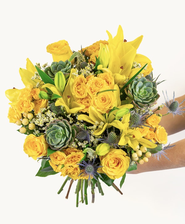 Vibrant bouquet of yellow flowers including roses and lilies with green succulents and foliage, held by hands against a white background.