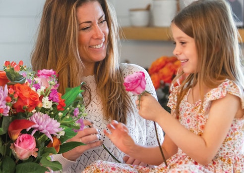 Smiling woman receiving a pink flower from a young girl, both sitting at a table with a colorful bouquet in a cozy, well-lit room, sharing a cheerful moment.