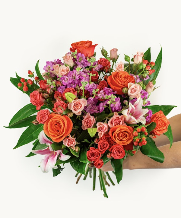 Hand holding a vibrant bouquet of flowers featuring orange roses, pink lilies, and purple accents with green foliage against a white background.