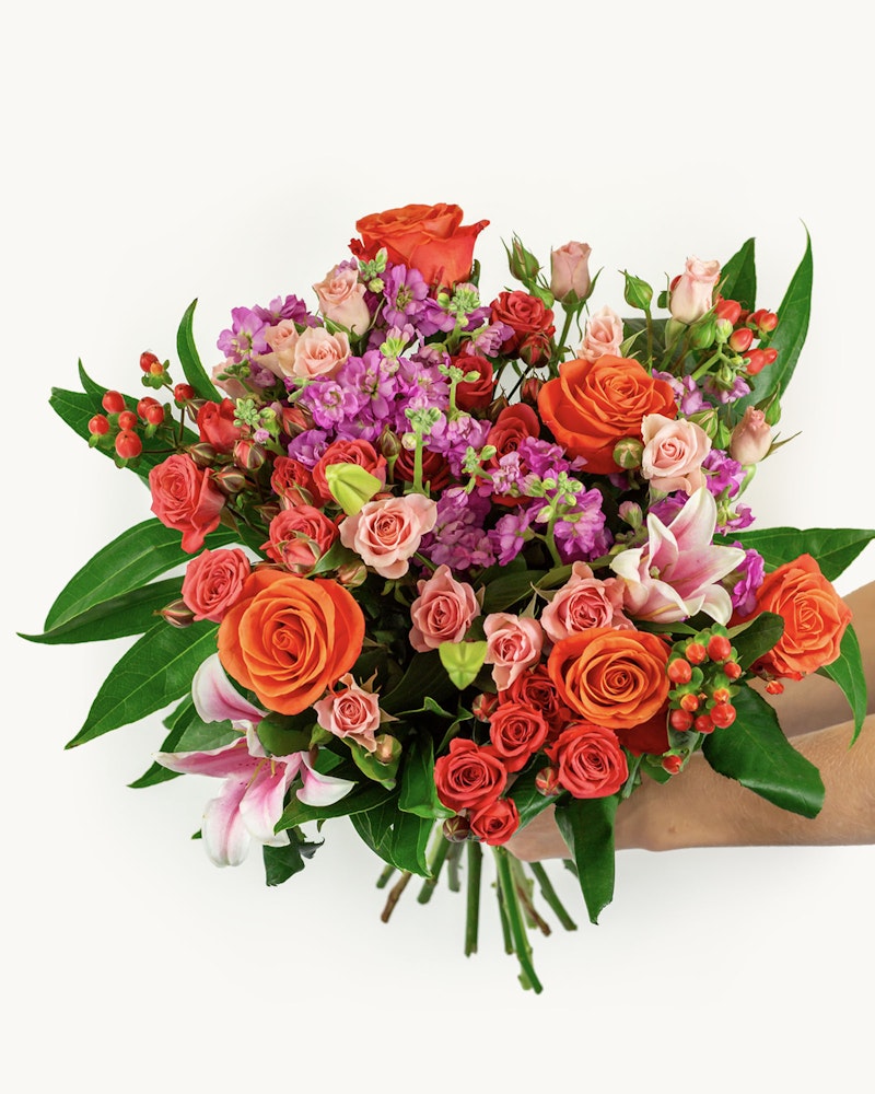 Hand holding a vibrant bouquet of flowers featuring orange roses, pink lilies, and purple accents with green foliage against a white background.