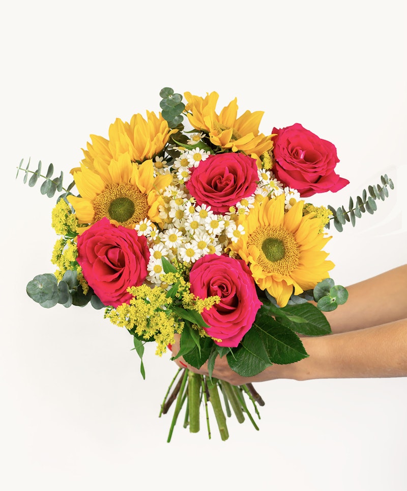 A vibrant bouquet of flowers with bright yellow sunflowers, deep pink roses, delicate white blossoms, and lush greenery held by a person against a white background.