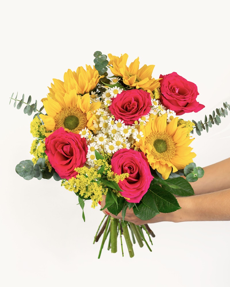 A vibrant bouquet of flowers with bright yellow sunflowers, deep pink roses, delicate white blossoms, and lush greenery held by a person against a white background.