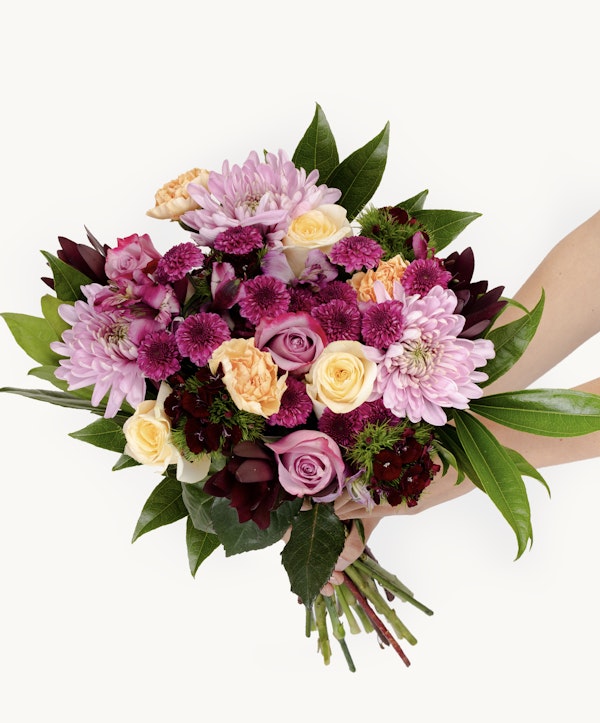 A person holds a vibrant bouquet featuring a mix of roses, chrysanthemums, and green foliage, with pink, purple, and orange hues against a white backdrop.