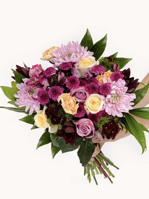 A person holds a vibrant bouquet featuring a mix of roses, chrysanthemums, and green foliage, with pink, purple, and orange hues against a white backdrop.
