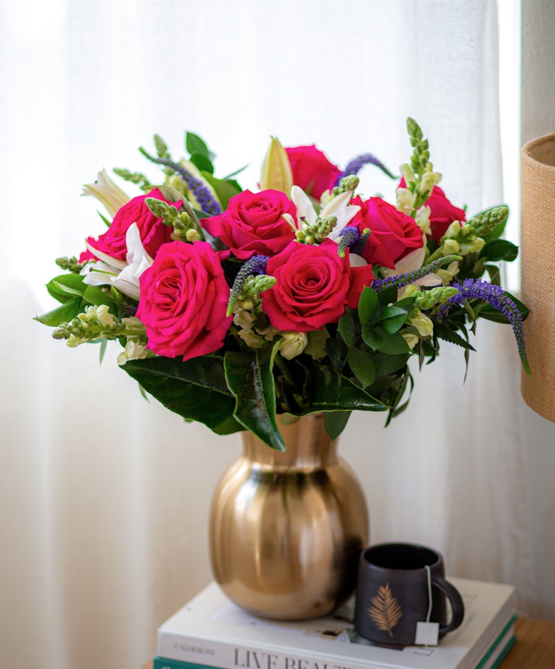 Vibrant bouquet of pink roses and white flowers arranged in a gold vase on a stack of books next to a window, with a decorative cup on a wooden surface.