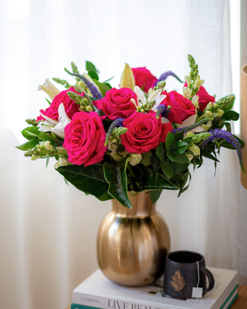 Vibrant bouquet of pink roses and white flowers arranged in a gold vase on a stack of books next to a window, with a decorative cup on a wooden surface.