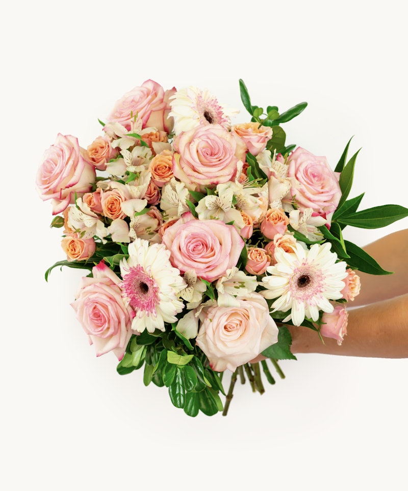 Overhead view of a vibrant bouquet featuring pink roses, light orange roses, white gerberas, and lush green leaves against a white background.