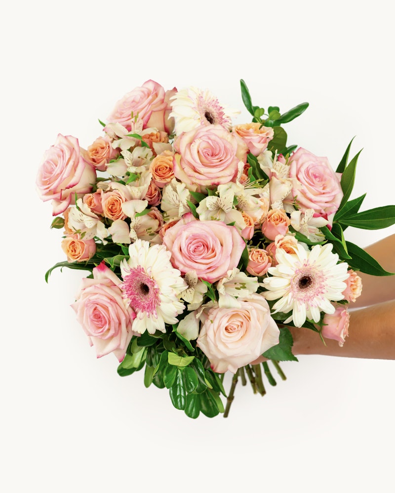 Overhead view of a vibrant bouquet featuring pink roses, light orange roses, white gerberas, and lush green leaves against a white background.