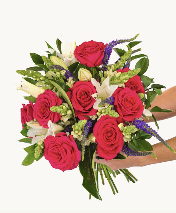 Vibrant bouquet of flowers featuring red roses, white lilies, and pink blooms with greenery and hints of purple flowers, held against a white background.